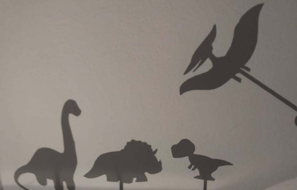 Brincadeiras de dinossauros com teatro de sombras: em uma parede, aparecem relfetidas formas imitando dinossauros (em pedaço de papel e palito)