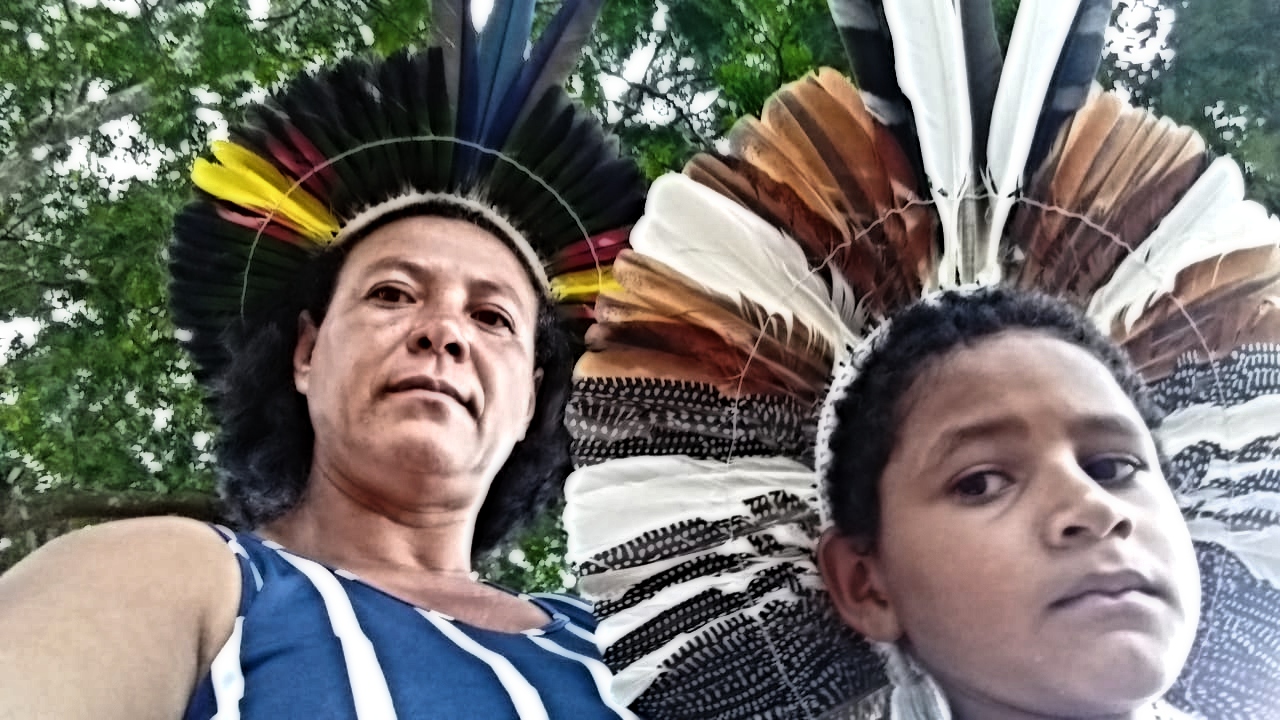 Uma mulher indígena do povo Xukuru-Kairiri, usando um cocar, está com seu filho à frente, também de cocar - ambos olham para o interlocutor (câmera)