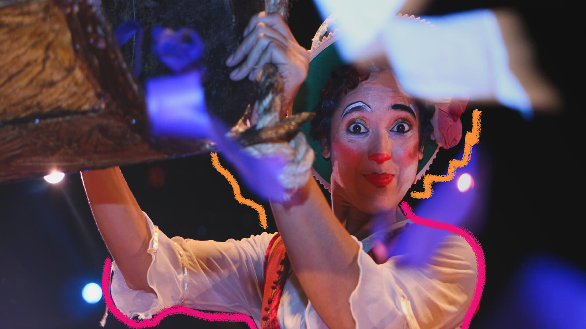 Na foto, artista circense vestida com blusa branca e adornada com pintura facial de palhaço aparece sorridente para a câmera