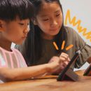 Conheça 5 aplicativos para aprimorar a aprendizagem infantil: na foto, duas crianças asiáticas olham para telas de tablet e celular. A imagem possui intervenções de rabiscos coloridos.