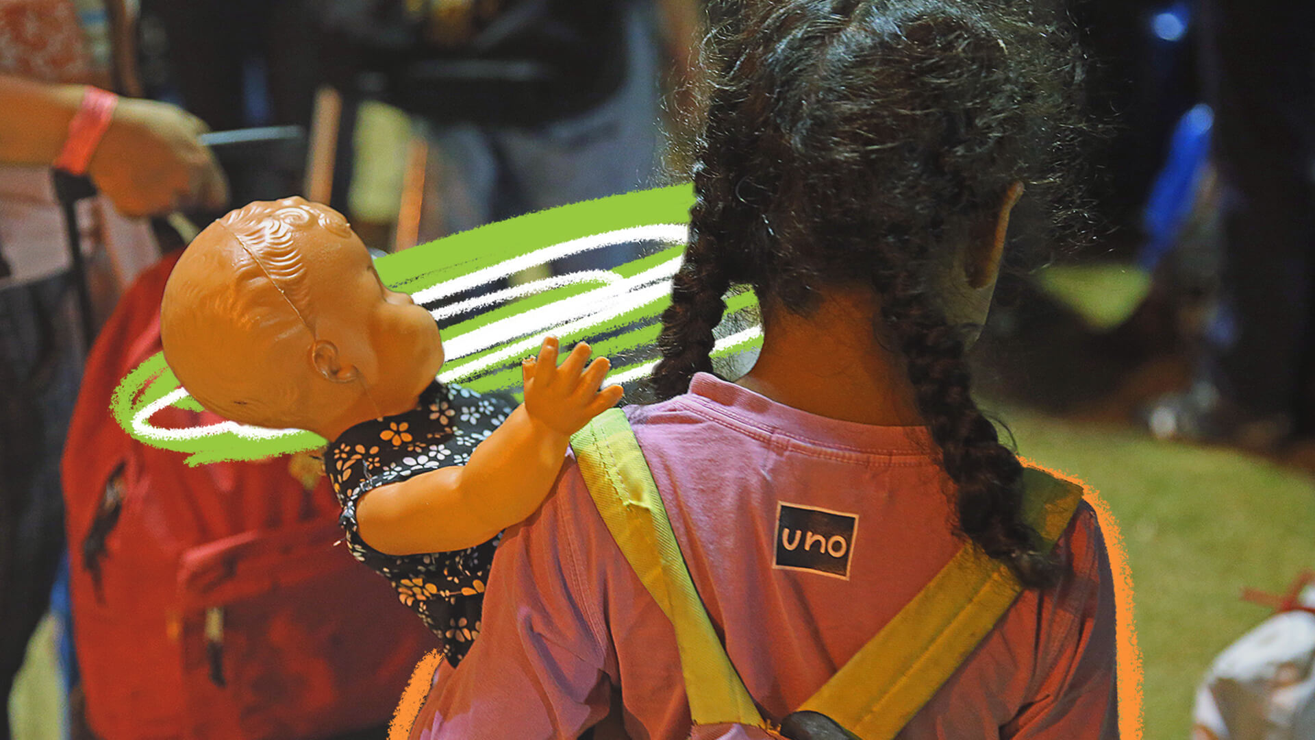 De costas, uma criança com cabelos negros trançados, usa camiseta rosa e uma mochila, e segura uma boneca de plástico com vestido florido no braço esquerdo.