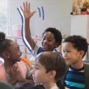 No chão da escola, Instituto Alana: quatro crianças, brancas e negras, estão em uma sala de aula. Um dos meninos está com o braço levantado