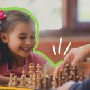 Uma menina de pele clara e cabelos lisos e negros, está sorrindo enquanto joga xadrez.