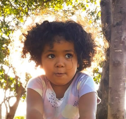 Uma menina negra de cabelos encaracolados, está posando para uma foto, mas com olhar direcionado para o lado