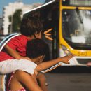 Foto de uma mulher, com uma criança sobre seus ombros, fazendo sinal para um ônibus parar