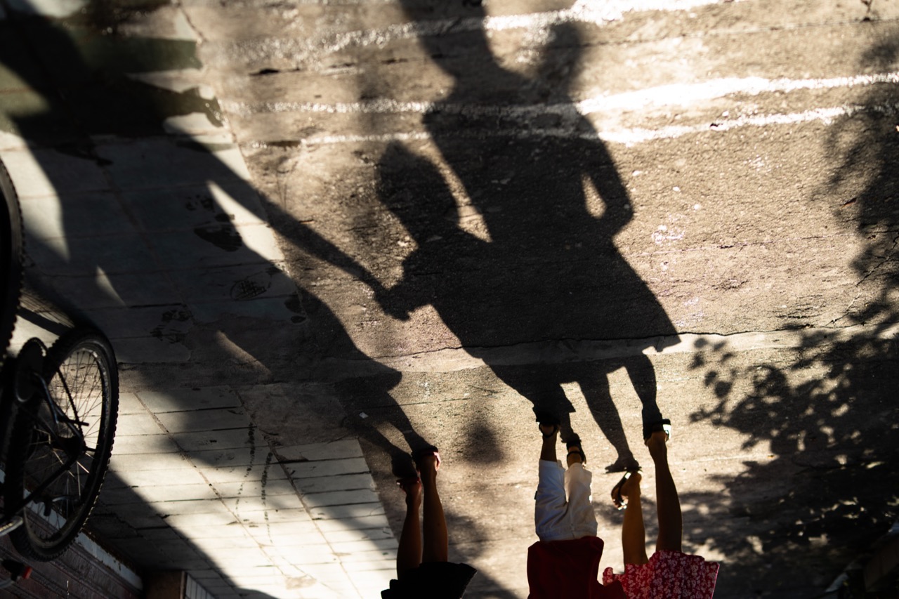 sombra projetada em uma calçada, de uma criança dando a mão para um adulto, e ao lado uma menina segurando uma boneca.