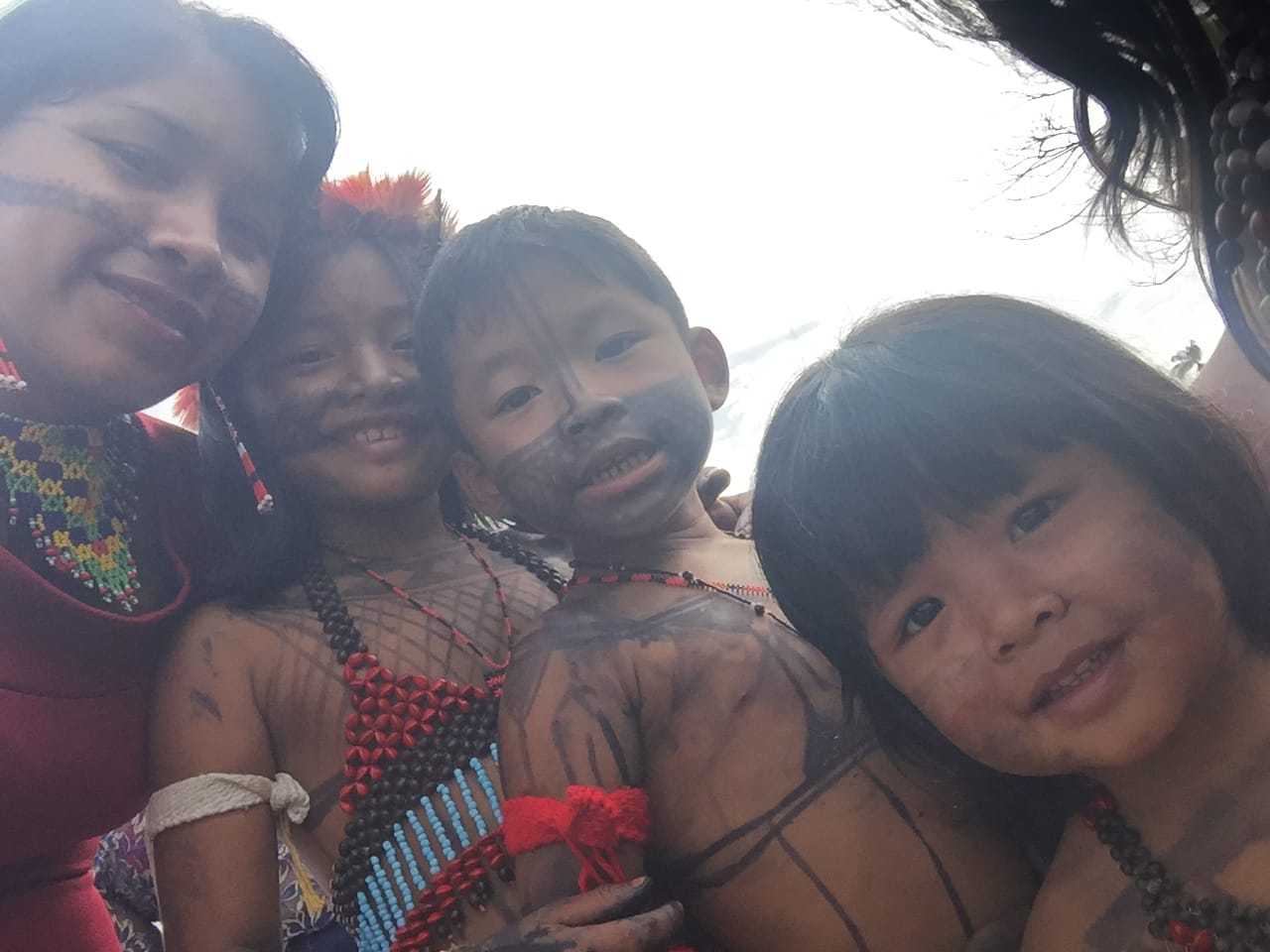  Selfie de uma mulher indígena, do lado direito, da foto, com três crianças indígenas, todas com pinturas espalhadas pelo rosto e corpo.