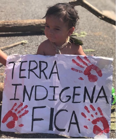 Foto de uma criança indígena segurando um cartaz onde se lê: “Terra indígena fica”