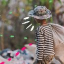 Foto de um menino de costas segurando um saco de tecido, sujo, enquanto caminha por um lixão. Ele veste calça bege, camisa de maga longa listrada de preto e branco e um chapéu com estampa militar.