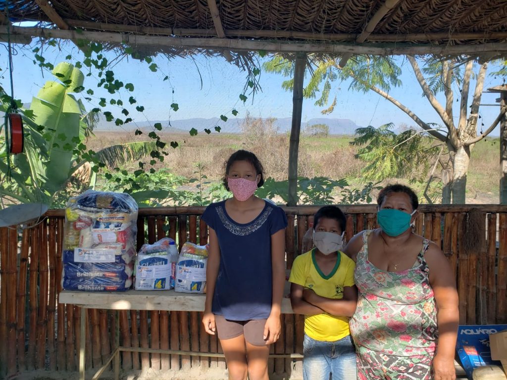 Uma menina (Naiara), está em pé ao lado de seu irmão menor e de sua mãe. Todos usam máscara de proteção respiratória. Ao fundo, está uma mesa com produtos de cesta básica e ao redor a mata devastada