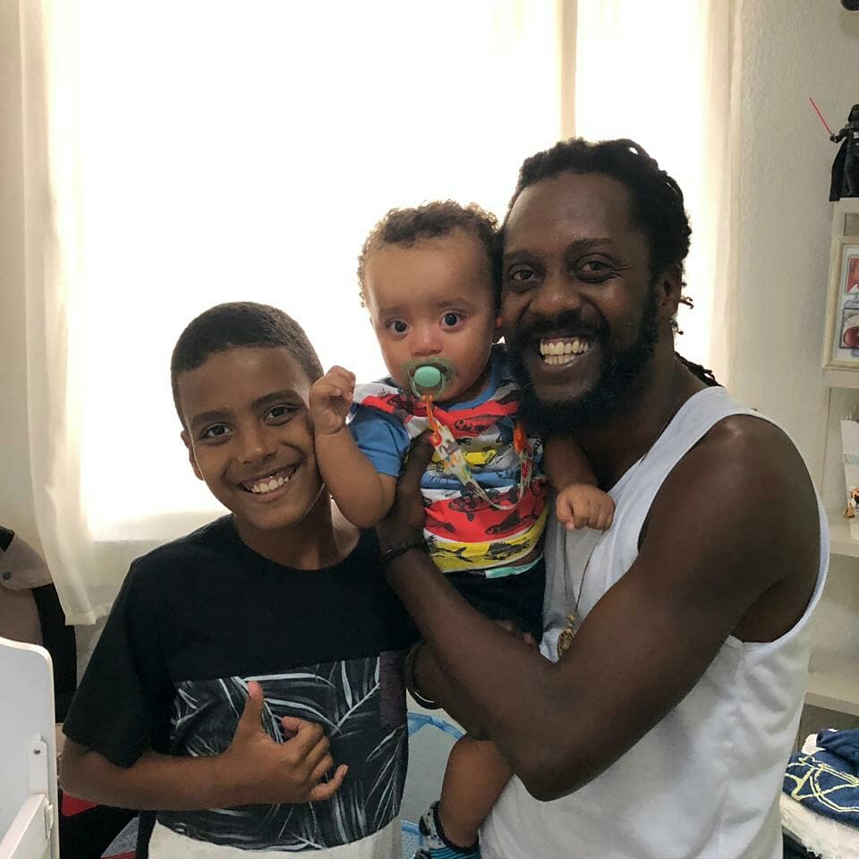 Um homem negro de regata branca, à direita na foto, sorri e segura um bebê também de pele negra com uma chupeta verde e camiseta colorida; ao lado do bebê, no canto esquerdo da foto, está um menino negro de camiseta preta, também sorrindo.