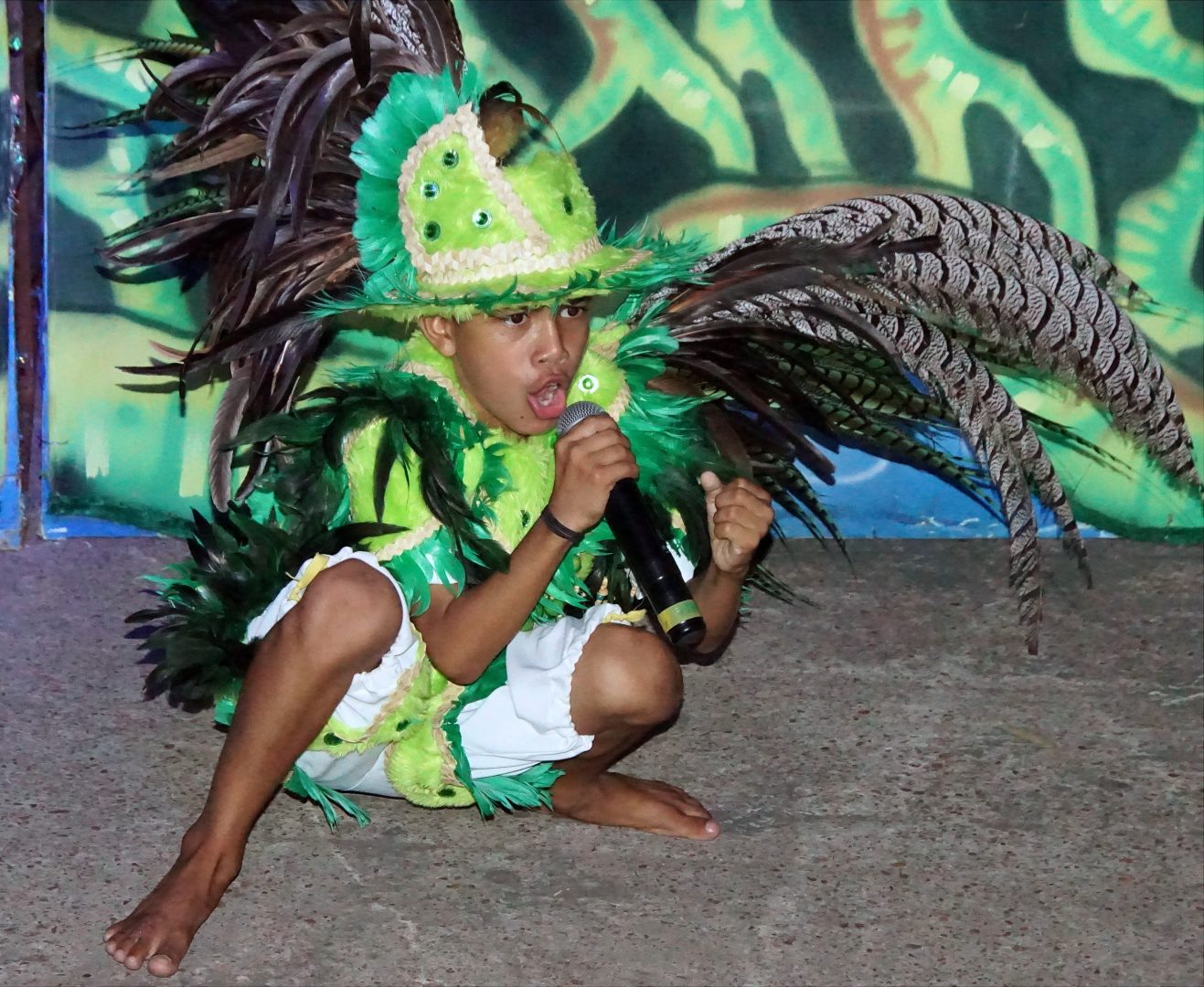 Uma criança com microfone na mão e agachada, usando chapéu e roupa verde com penas