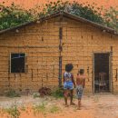 Foto de duas crianças negras, de costas, andando em direção a uma casa feita de barro