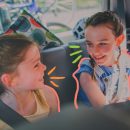 Foto de duas meninas sentadas no banco de detrás do carro, usando cinto de segurança. Elas conversam e se divertem com brincadeiras enquanto esperam a viagem terminar.