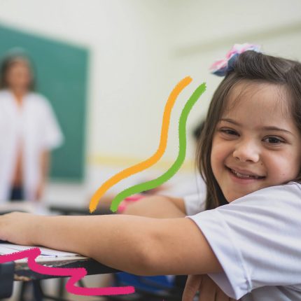 Inclusão na escola: foto de uma menina está sentada em sua carteira na sala de aula. Ao fundo, a professora aparece desfocada em frente à lousa.