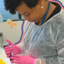 Foto de Jaqueline Goes, uma cientista negra brasileira. Ela está num laboratório, usa roupa e luvas de proteção, enquanto manipula testes clínicos.