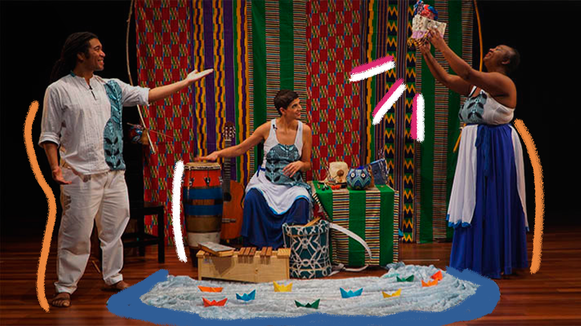 Cena da peça Canto das águas. Um homem e duas mulheres estão num cenário com elementos de origem africana e, no meio, entre eles, há um rio com barquinhos coloridos de papel