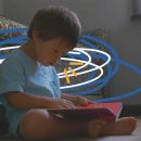 Cena do filme "Um lugar para todo mundo" em que Emílio, um menino que tem síndrome de Down, está sentado no chão, encostado no sofá, com um livro vermelho aberto no colo. Ele veste uma camiseta azul.