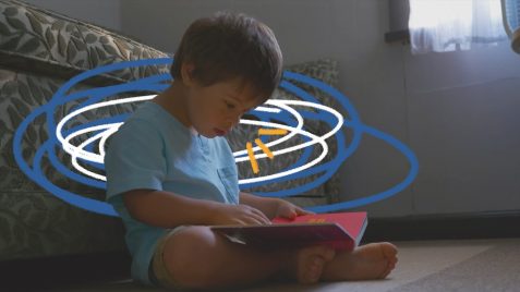Cena do filme "Um lugar para todo mundo" em que Emílio, um menino que tem síndrome de Down, está sentado no chão, encostado no sofá, com um livro vermelho aberto no colo. Ele veste uma camiseta azul.