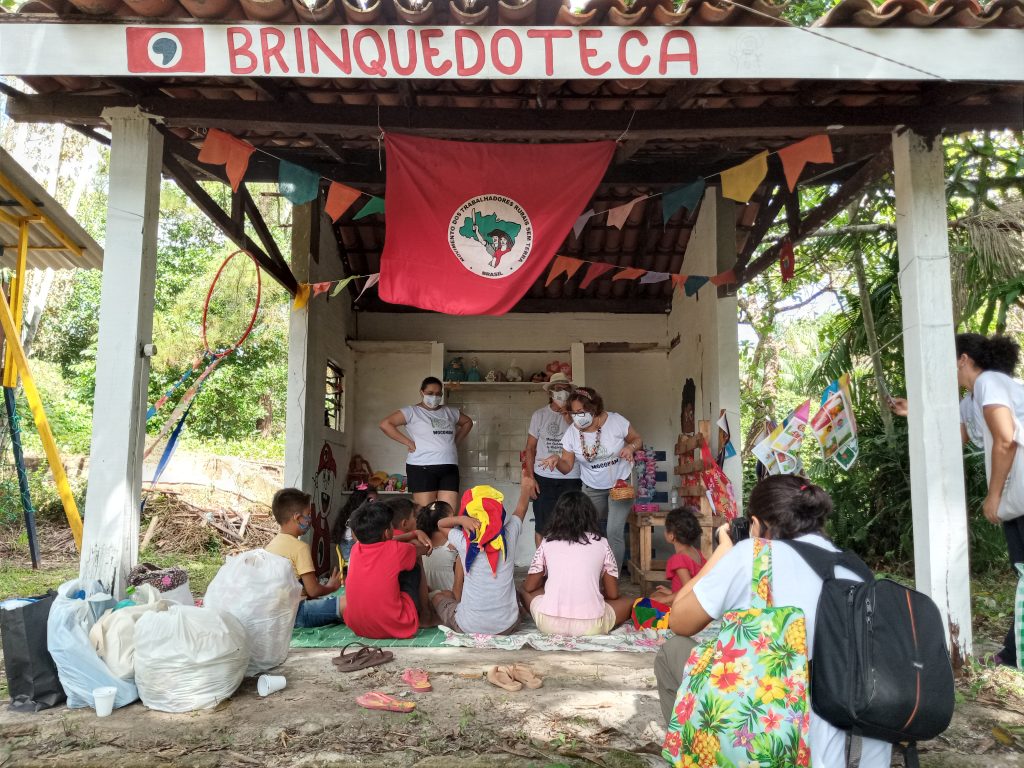 Foto de crianças sentadas no chão de terra batida, ouvindo três adultos, embaixo de um local coberto, mas sem paredes nem portas. A bandeira do MST está no teto, onde se lê também “Brinquedoteca”.