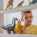 Na foto, um menino de pele clara e cabelos loiros brinca com miniaturas de dinossauros em uma estante. A imagem possui intervenções coloridas nas cores laranja, verde e azul.