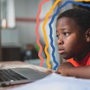 Na foto, um menino negro encara a tela de um computador, que está junto de folhas de papel