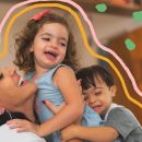 Uma mulher abraça duas crianças. A menina tem cabelos crespos com um laço rosa e vestido azul. O menino veste camiseta cinza e tem síndrome de Down.