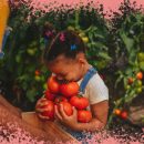 Na foto, uma menina negra sorri enquanto segura vários tomates. A imagem contém uma intervenção de moldura na cor rosa.
