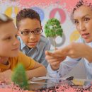 Na foto, dois meninos olham para uma árvore em miniatura, mostrada por uma professora. A imagem possui uma intervenção de moldura na cor rosa.