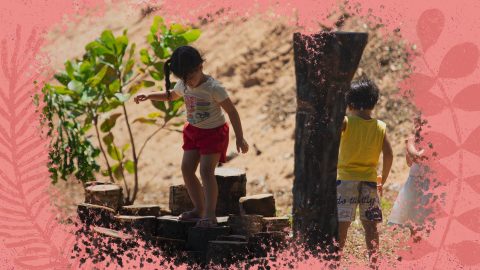 Na foto, duas crianças brincam entre troncos de árvores. A imagem possui uma intervenção de moldura na cor rosa.