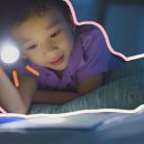 Foto de um menino deitado na cama de bruços sobre um livro. Ele lê no escuro com a ajuda de uma lanterna.