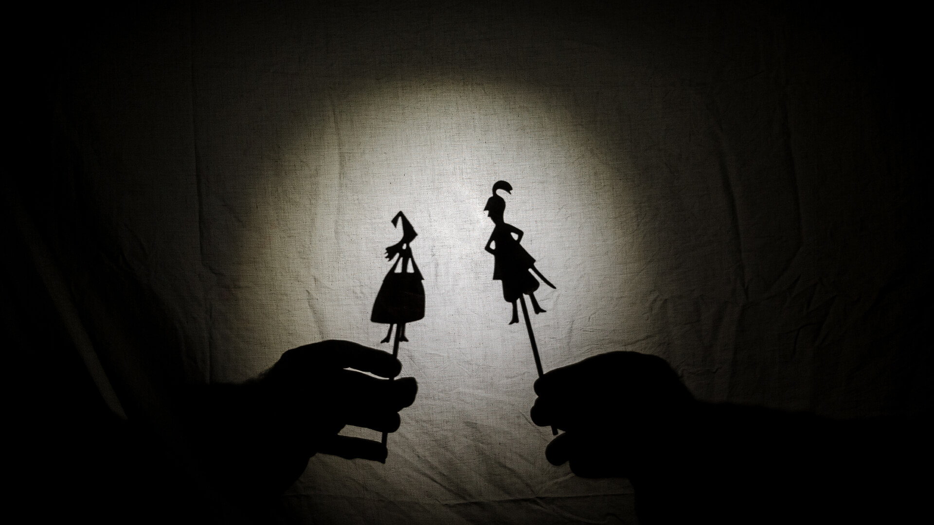 Duas mãos seguram personagens recortadas de papel num teatro de sombras