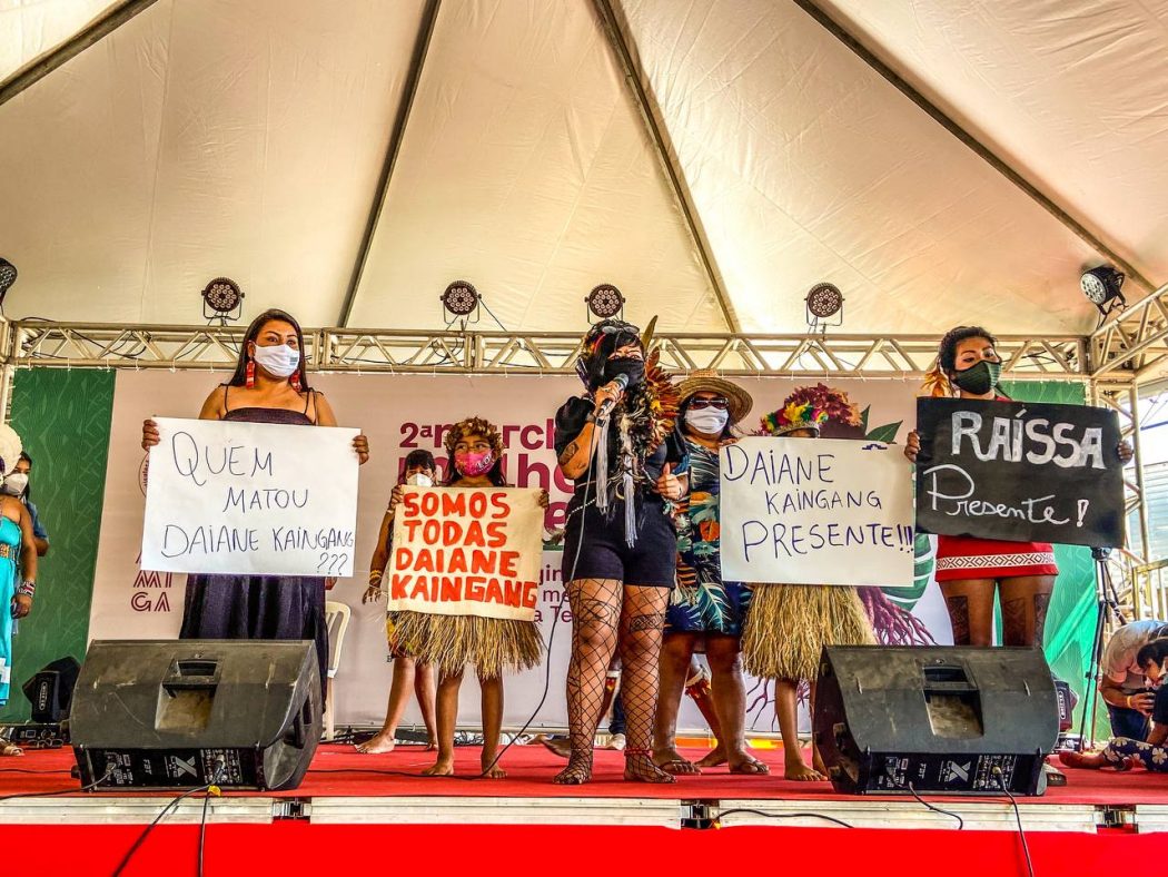 Mulheres indígenas em cima de um palco segurando cartazes, onde se lê, por exemplo, "Raissa presente" e "Quem matou Daiane Kaingang?"