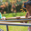 Foto de duas crianças negras, um menino e uma menina, num brinquedo de girar em um parque ao ar livre. Ele veste camiseta verde e ela uma regata rosa e uma tiara de flor azul no cabelo