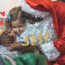 Na foto, uma menina recebe o abraço de um Papai Noel. A imagem possui intervenções de rabiscos coloridos.