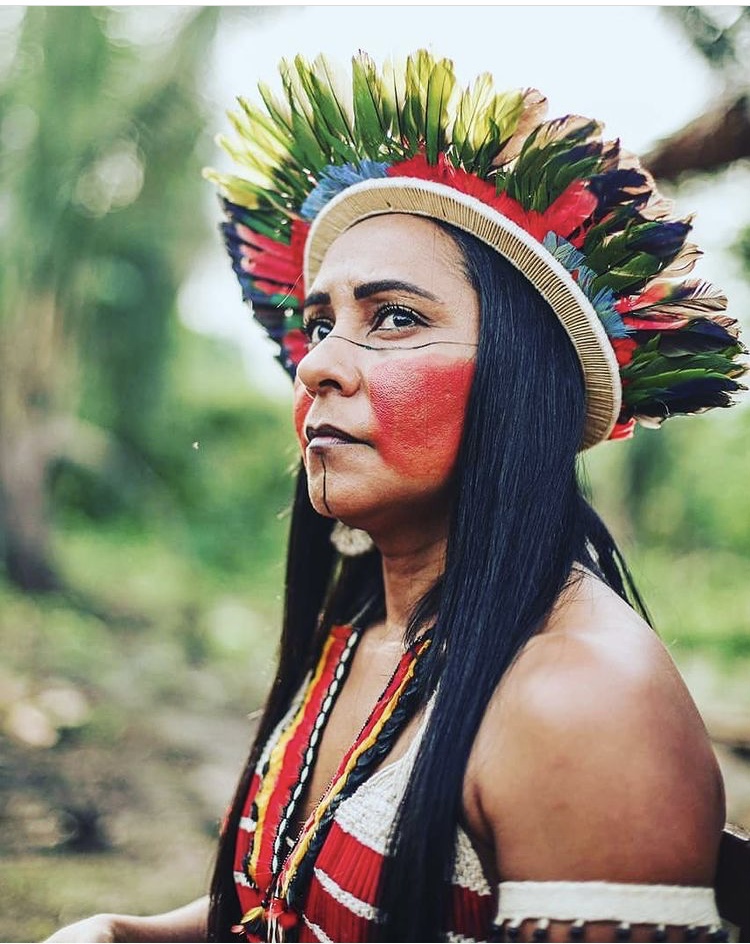 Uma mulher indígena com adereços e pintura tradicionais olha para o horizonte. Ao fundo, árvores e folhagens
