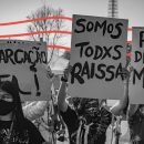 Foto em preto e branco de uma manifestação de rua em que pessoas levam cartazes onde se lê, por exemplo, "Somos todos Raissa", "Demarcação já" e "Pare de nos matar". Há intervenções de arte com linhas vermelhas