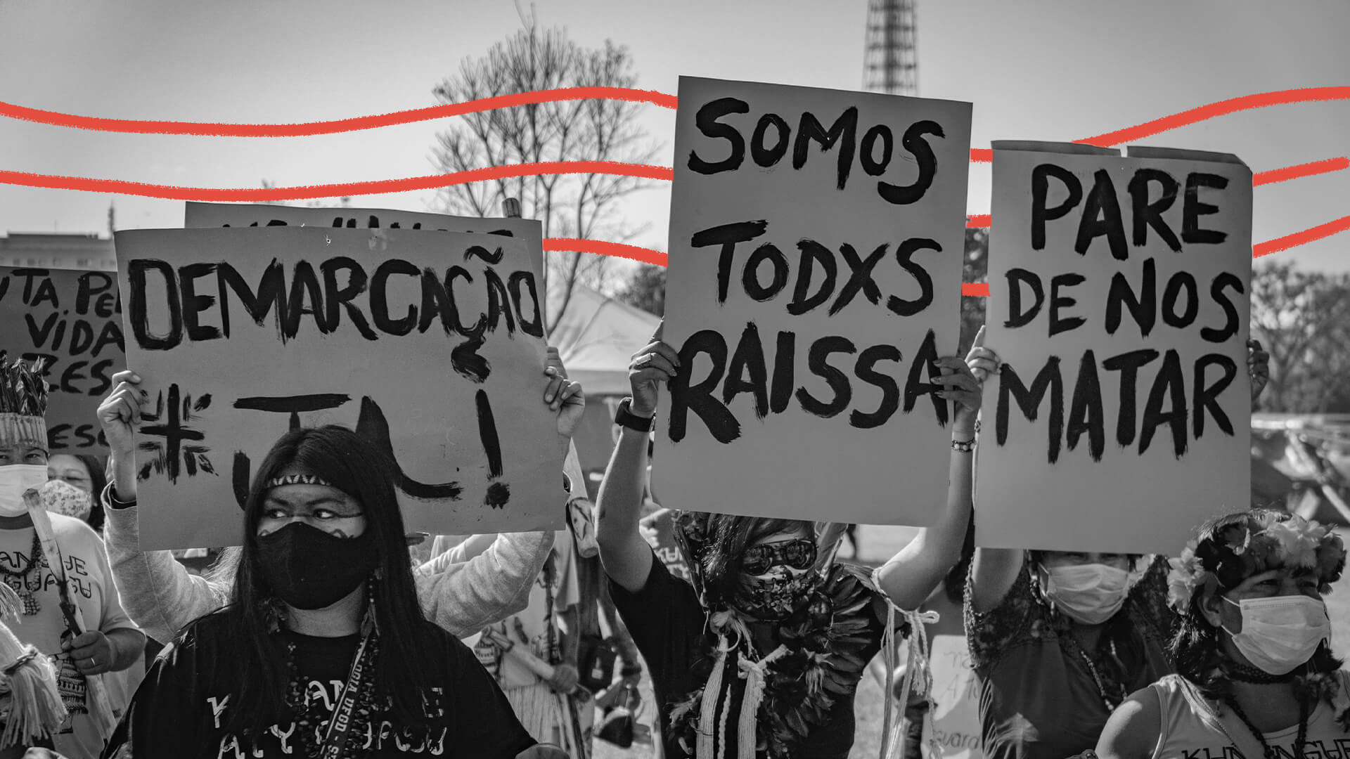 Foto em preto e branco de uma manifestação de rua em que pessoas levam cartazes onde se lê, por exemplo, "Somos todos Raissa", "Demarcação já" e "Pare de nos matar". Há intervenções de arte com linhas vermelhas