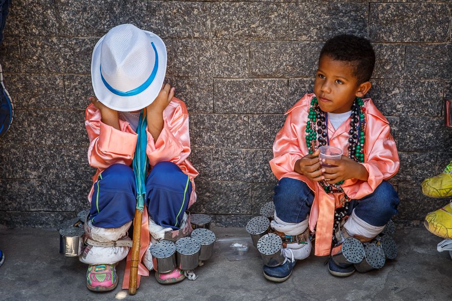 Dois meninos estão vestidos para a festa do Congado. Ambos estão agachados contra uma parede de pedra, e um deles tem o rosto escondido pelo chapéu