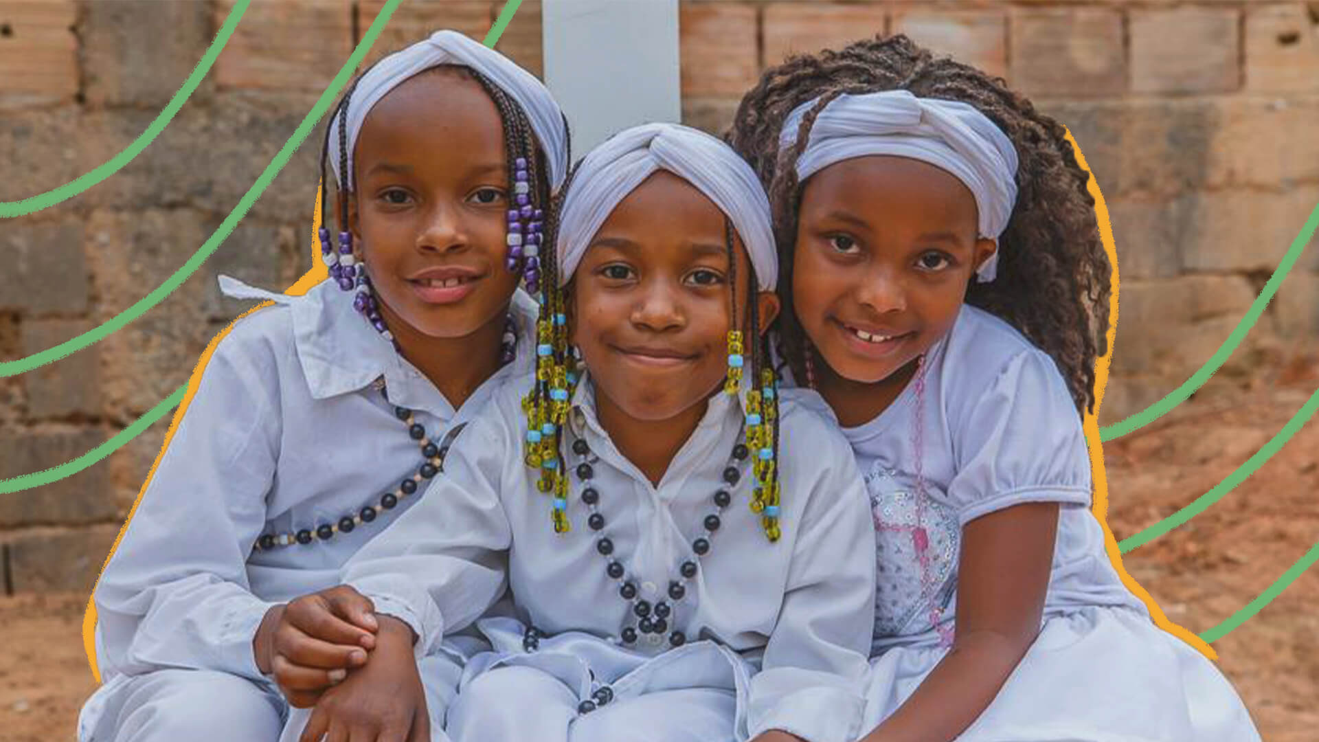 Três crianças negras vestidas com roupas brancas e colares de conta estão sentadas e de mãos dadas, sorrindo para a foto