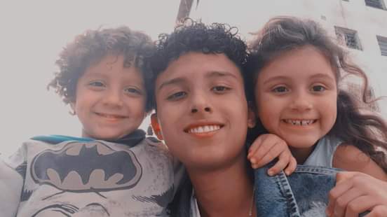 Três crianças posam para a foto com as rostos próximos e sorrisos