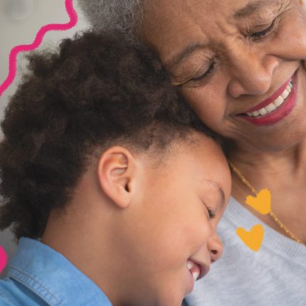 Avó e neto se abraçam e sorriem. Ela é uma mulher negra e grisalha, de cabelos curtos. Ele tem o cabelo cacheado e usa camisa jeans. Os dois estão sentados num sofá.