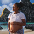Uma mulher grávida está na praia. Ela olha para o lado e apoia as duas mãos na barriga. A matéria é sobre bebês que não podem nascer em Fernando de Noronha.