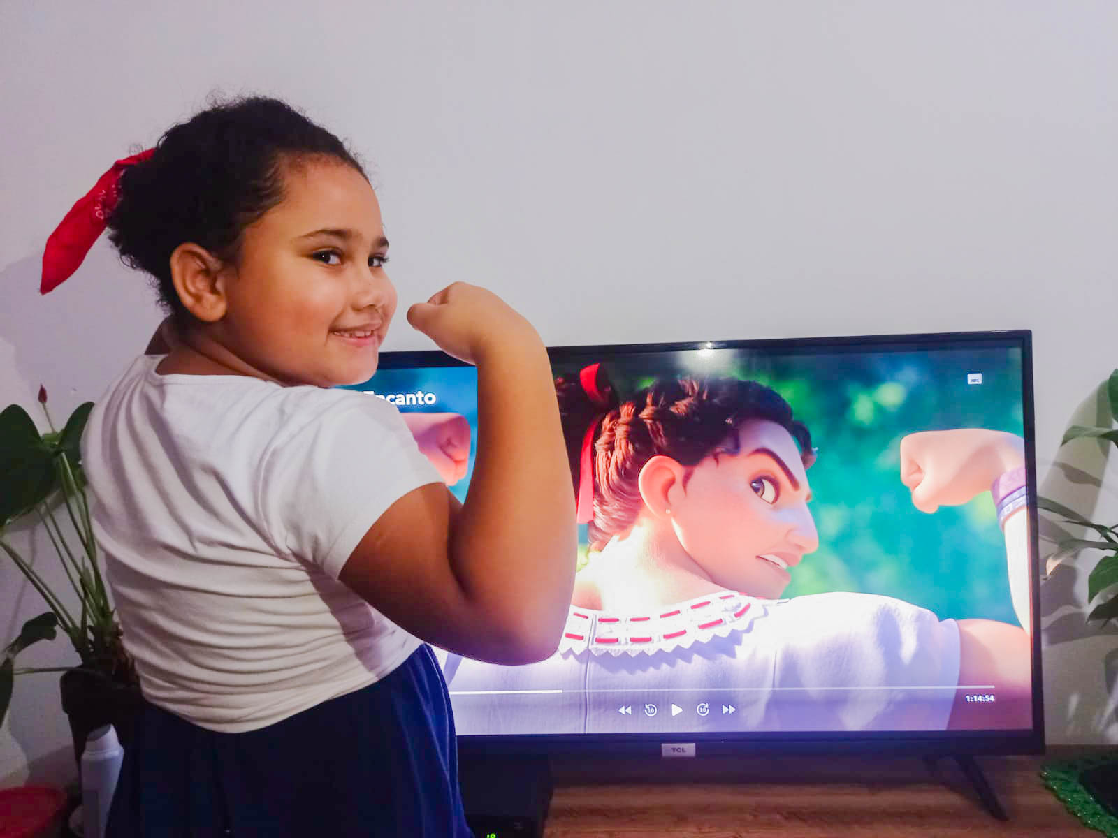 Uma menina de coque faz o mesmo gesto com o braço que a personagem Luisa, do filme "Encanto", que está na tela da televisão