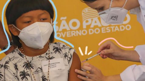 Foto com funda amarelo mostra menino indígena de máscara, em primeiro plano, recebe vacina aplicada no braço por profissional de saúde.