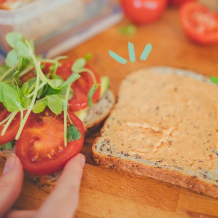 Na imagem, mãos de pele clara preparam um sanduíche. A foto mostra duas fatias de pão recheadas com uma pasta na cor laranja, tomate e agrião. A imagem possui intervenções de rabiscos coloridos.