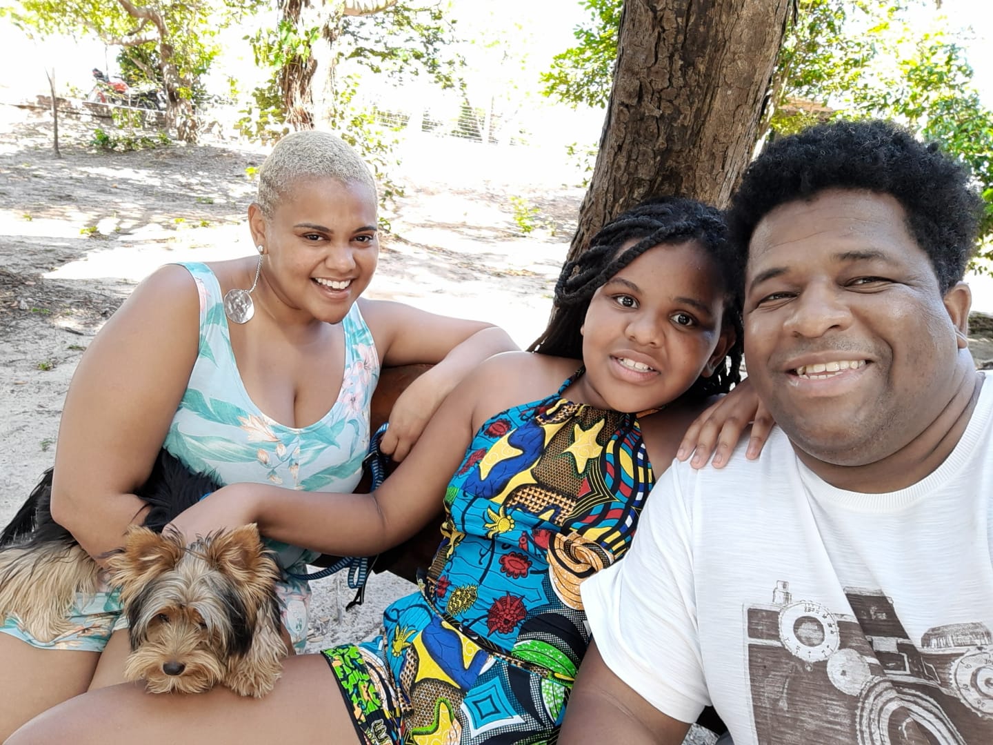 Uma família de pessoas negras composta por uma mulher, um homem e uma menina. Eles estão num espaço ao ar livre e a criança tem um cachorro no colo. Todos sorriem para a foto
