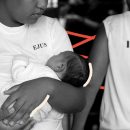 Como fica o direito à prisão domiciliar de mães encarceradas?. Imagem em preto e branco mostra uma mãe, negra, segurando um bebê no colo. A foto possui intervenções de rabiscos coloridos na cor vermelha.