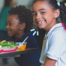 Projeto ‘Comida de verdade’: autonomia alimentar começa na escola: na imagem, duas crianças estão sentadas frente a uma mesa. O destaque é em uma menina, negra, que sorri com um prato de comida ao seu lado. No fundo da imagem, um menino negro também sorri. A imagem possui intervenções de rabiscos coloridos.