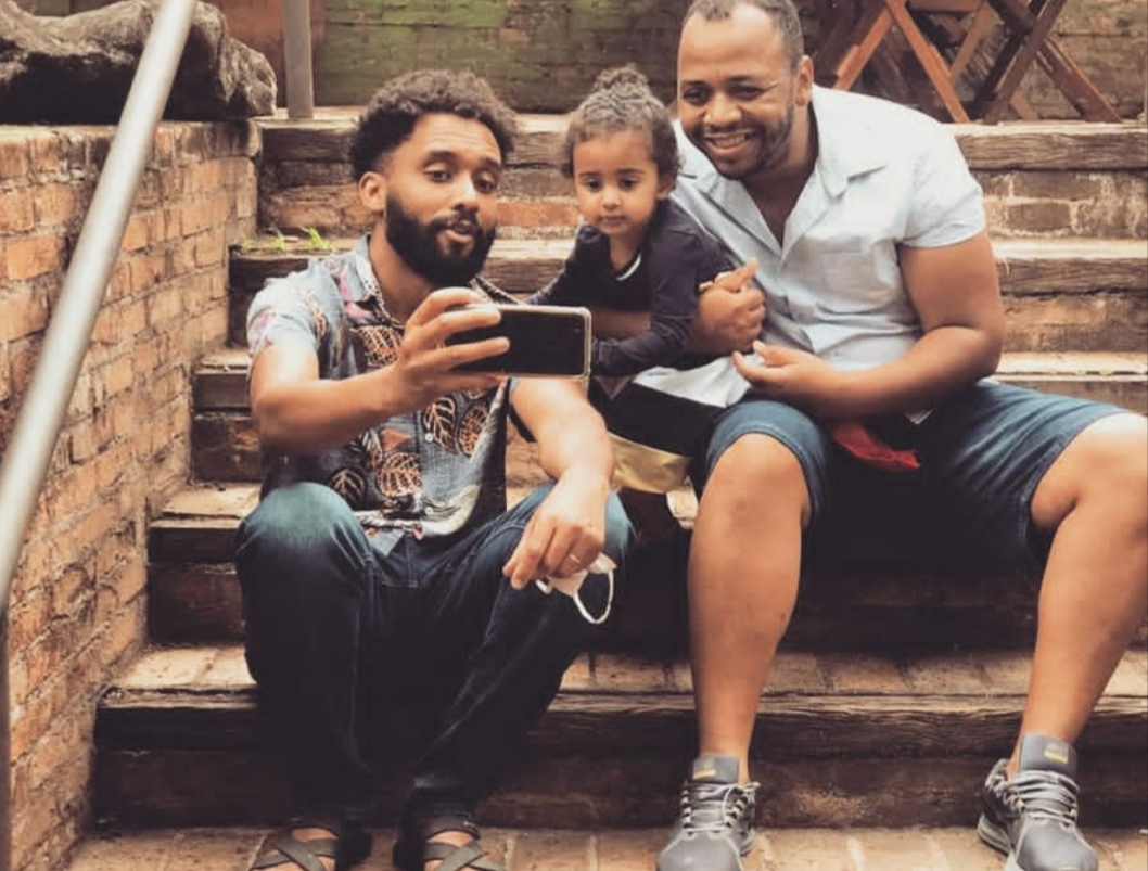 Um casal composto por dois homens tira uma selfie com a filha pequena. Os três estão sentados numa escadaria e são negros
